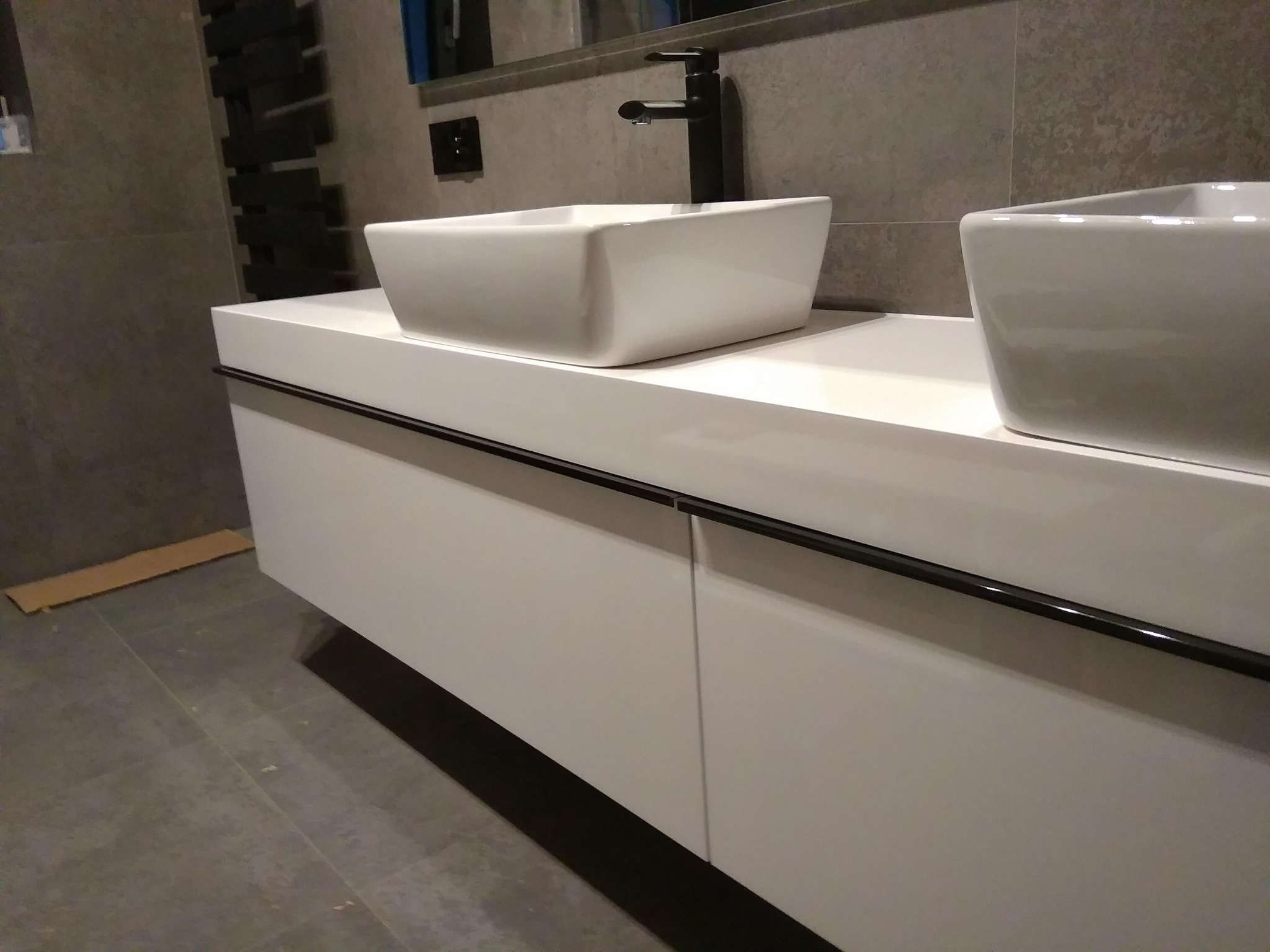 Nowoczesna łazienka z białą szafką pod umywalki nablatowe i czarną armaturą, szara płytka na ścianie i podłodze, widoczny fragment grzejnika drabinkowego.