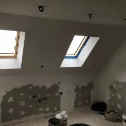 Pokój na poddaszu w trakcie remontu: ściany częściowo pokryte szpachlą, widoczne otwory na gniazdka elektryczne, dwa okna dachowe, puszki z farbą i narzędzia na podłodze.