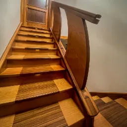 Drewniane schody z dwukolorowymi stopniami w szachownicę, podświetlane, z zakrzywioną balustradą i dekoracyjnymi elementami, prowadzące do drewnianych drzwi.