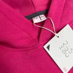 Różowa bluza z kapturem z metką marki Hajducka, prezentująca logo i elementy identyfikacji wizualnej.