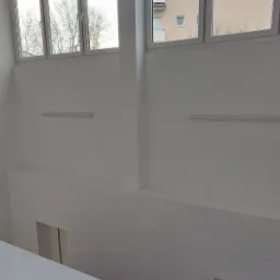 Wnętrze w trakcie prac wykończeniowych: białe ściany z oknami, surowe betonowe schody prowadzące w dół, widoczne lampy sufitowe.