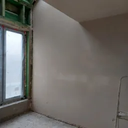 Świeżo otynkowane ściany w pomieszczeniu z oknem zabezpieczonym folią, widoczna drabina malarska i surowe wykończenie ramy okiennej.