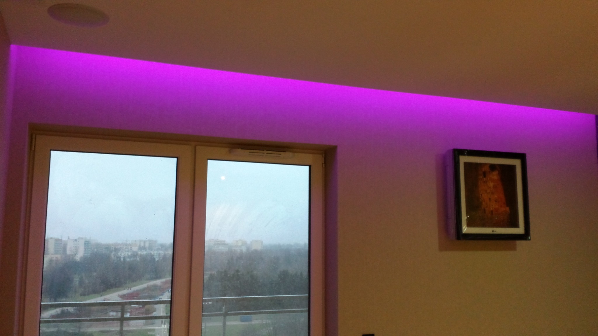 Pokój z podświetleniem LED w kolorze fioletowym nad oknem balkonowym i obrazem Gustava Klimta na ścianie.
