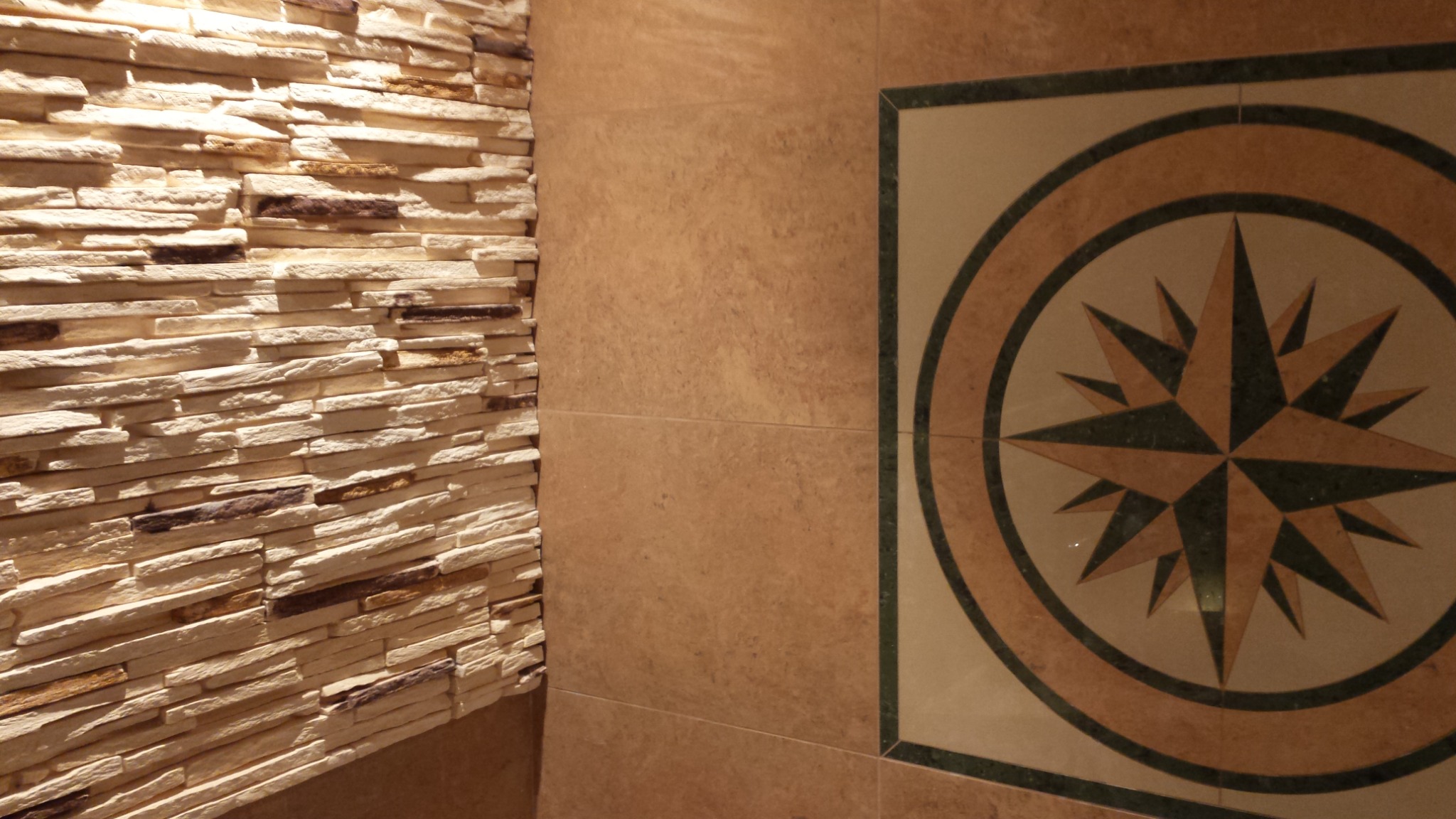 Ściana wyłożona dekoracyjnym kamieniem imitującym piaskowiec, sąsiadująca z gładką, brązową ścianą i fragmentem podłogi z motywem kompasu z zielonymi akcentami.
