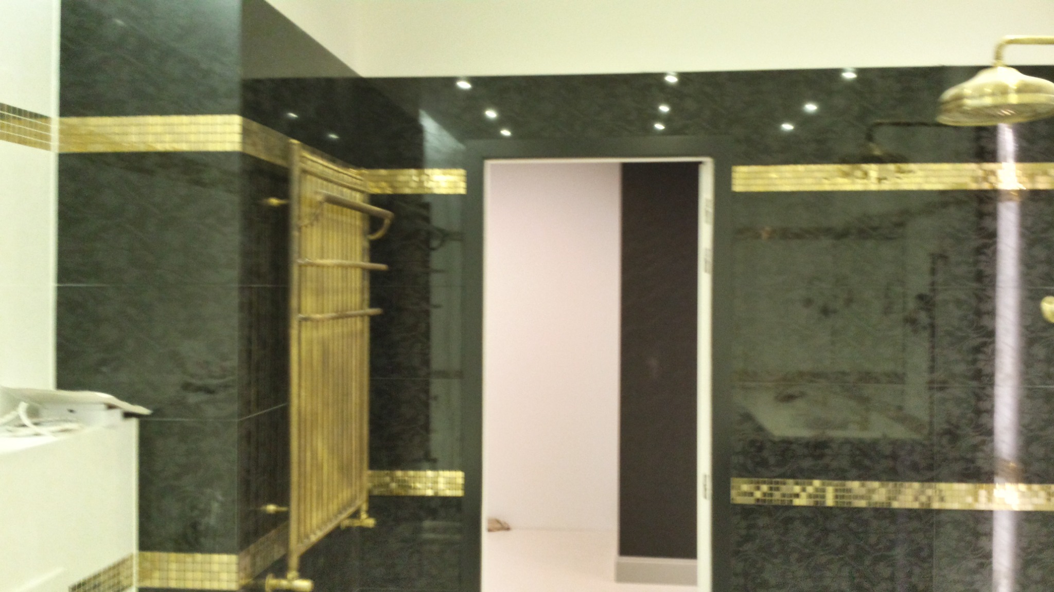 Elegancka łazienka z ciemnozielonymi płytkami, złotymi akcentami i grzejnikiem drabinkowym w stylu retro. Widoczny prysznic deszczownica i oświetlenie punktowe.