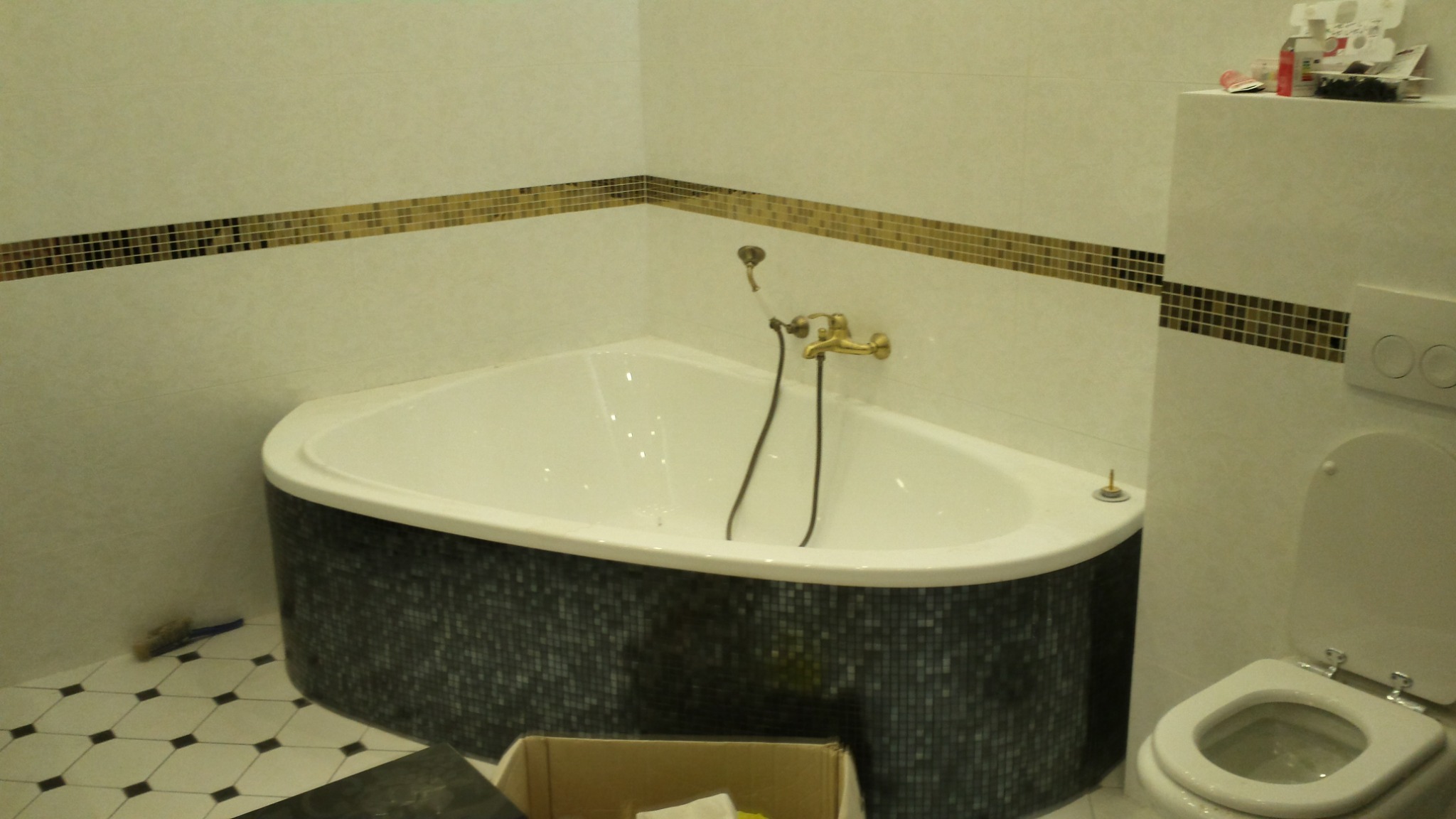 Narożna wanna obudowana mozaiką w wyremontowanej łazience, z widoczną armaturą w stylu retro i dekoracyjnym pasem mozaiki na ścianach, obok toaleta i niedbale pozostawione akcesoria.