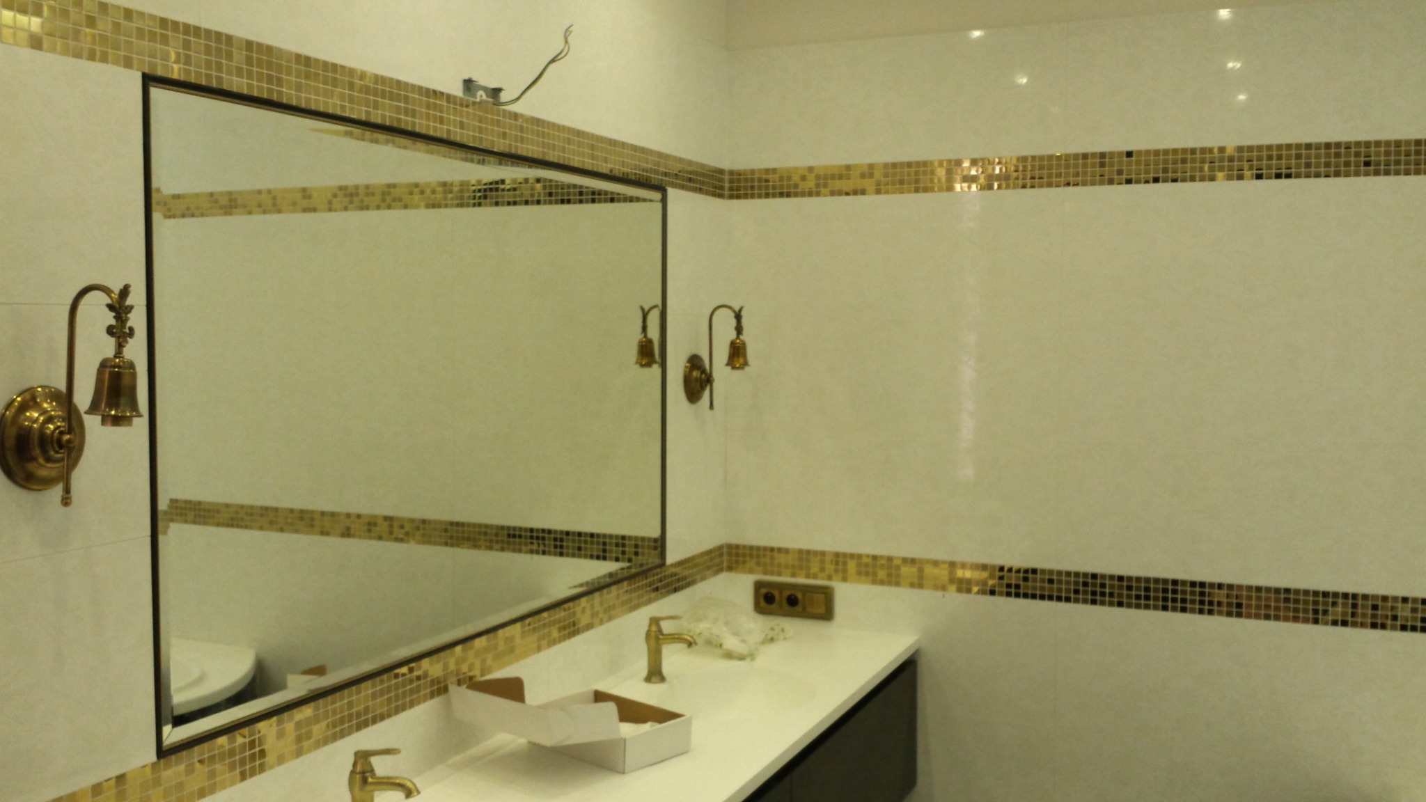 Wykończona łazienka z białymi płytkami i dekoracyjnym pasem z mozaiki w odcieniach złota, duże lustro z czarną ramą i złotym obramowaniem z mozaiki, złote kinkiety w stylu retro, biała umywalka...