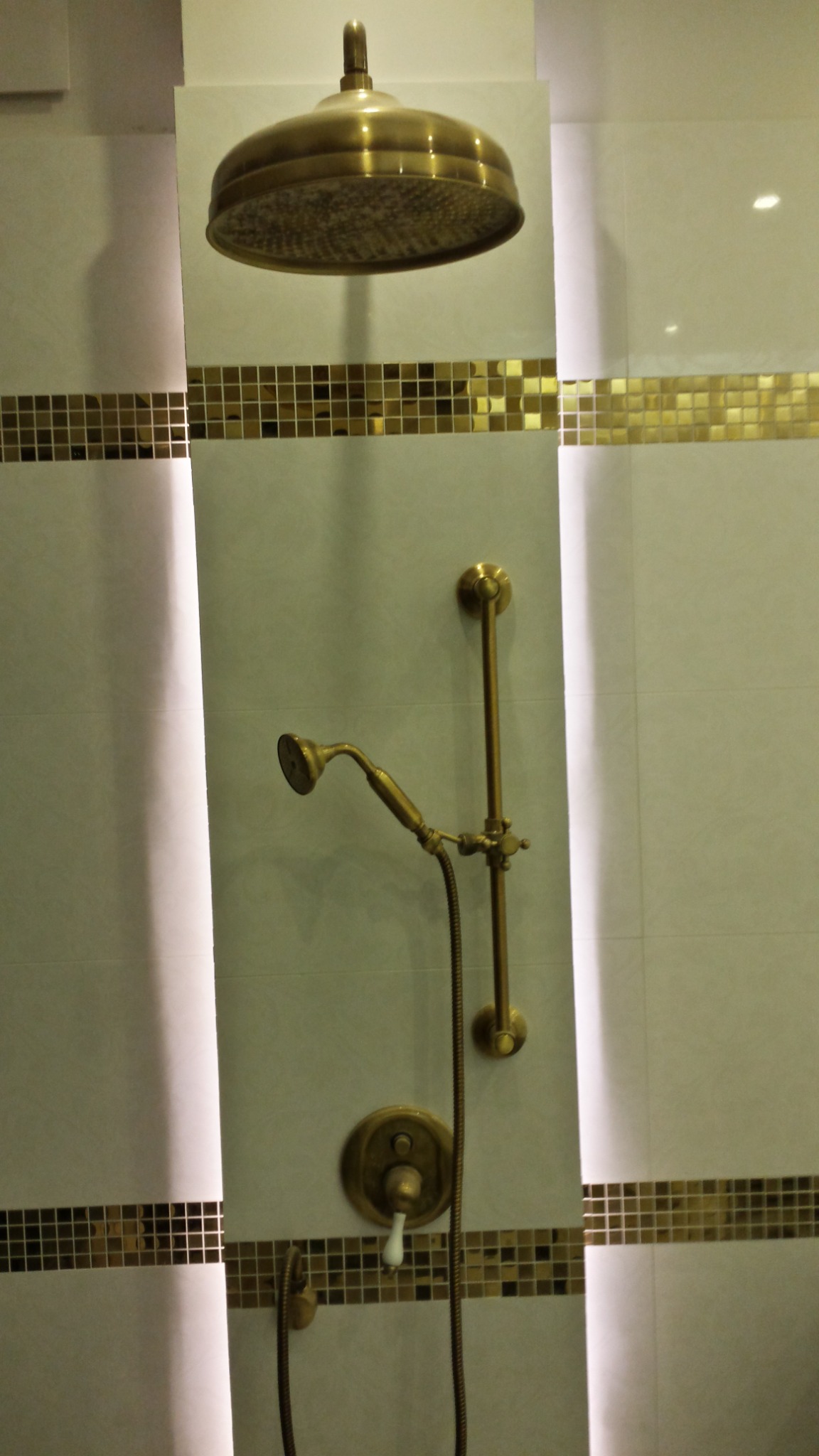 Wykończona łazienka z armaturą w stylu retro: deszczownica, słuchawka prysznicowa i bateria w kolorze starego złota, białe płytki z poziomym pasem złotych mozaiek.