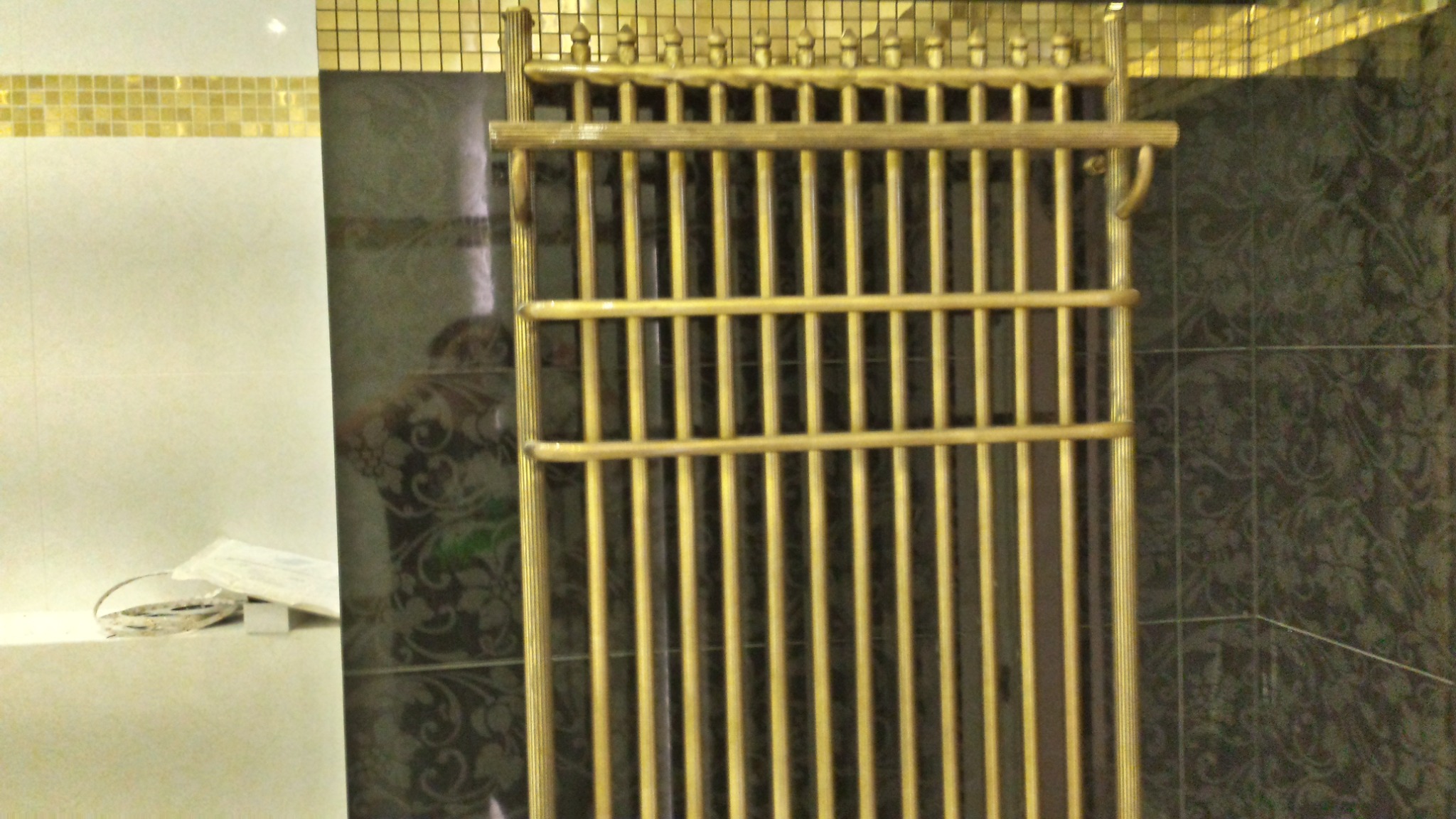 Złoty grzejnik łazienkowy drabinkowy na tle płytek z motywem florystycznym, widoczne fragmenty białych płytek z mozaiką.