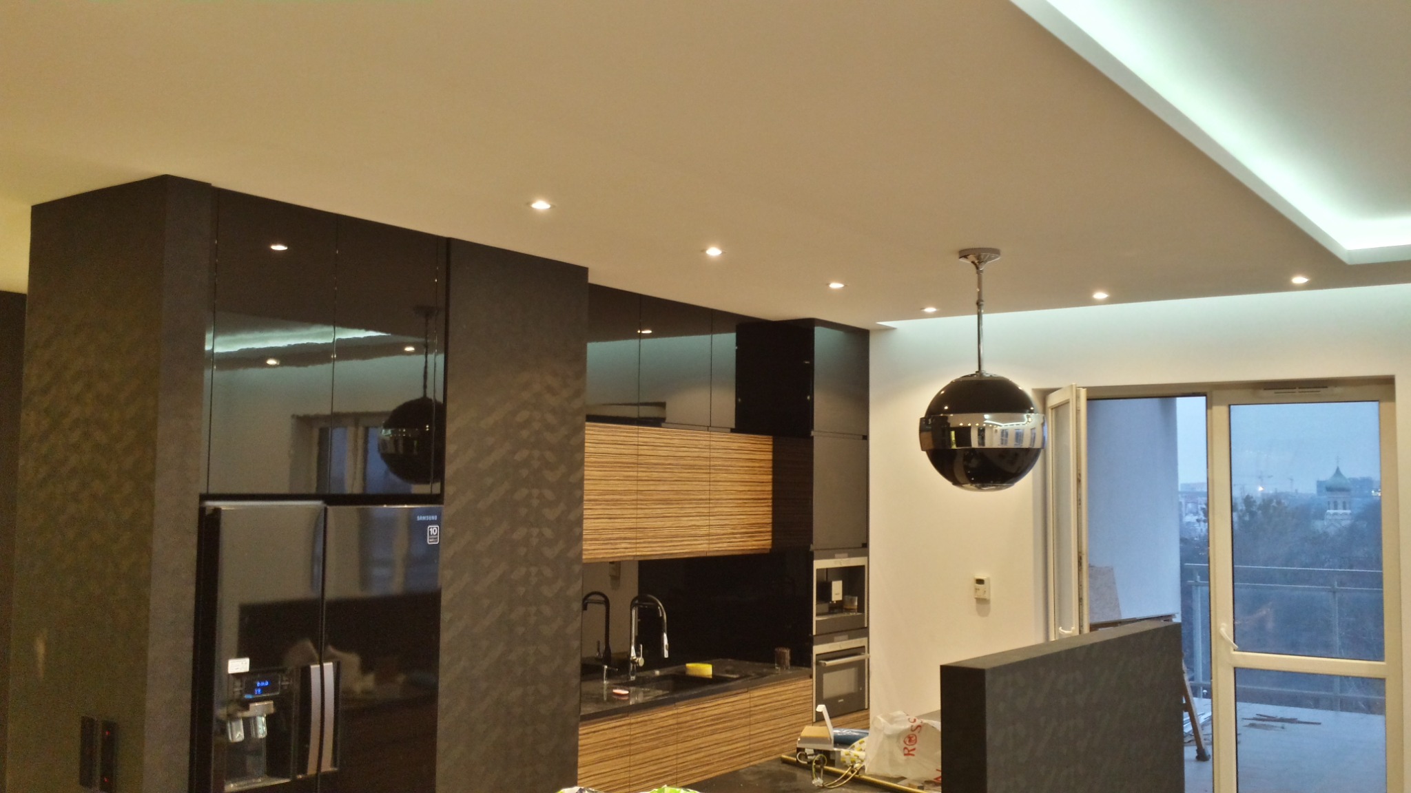 Nowoczesna kuchnia z czarnymi, lakierowanymi szafkami, drewnianymi akcentami i kulistą lampą wiszącą; widok na miasto przez otwarte drzwi balkonowe.