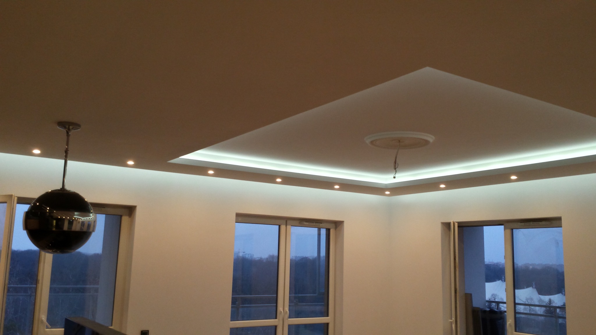 Nowoczesny salon z podwieszanym sufitem z oświetleniem punktowym i paskiem LED, widok na zewnątrz przez okna balkonowe.