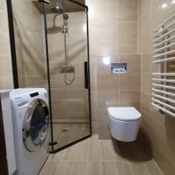 Nowoczesna łazienka z kabiną prysznicową typu walk-in z czarną ramą, podwieszaną toaletą, grzejnikiem drabinkowym i pralką Candy, wykończona beżowymi płytkami imitującymi drewno.