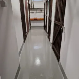 Korytarz z rzędem drewnianych boksów z kłódkami, szare ściany i podłoga z widoczną instalacją wodno-kanalizacyjną pod sufitem.