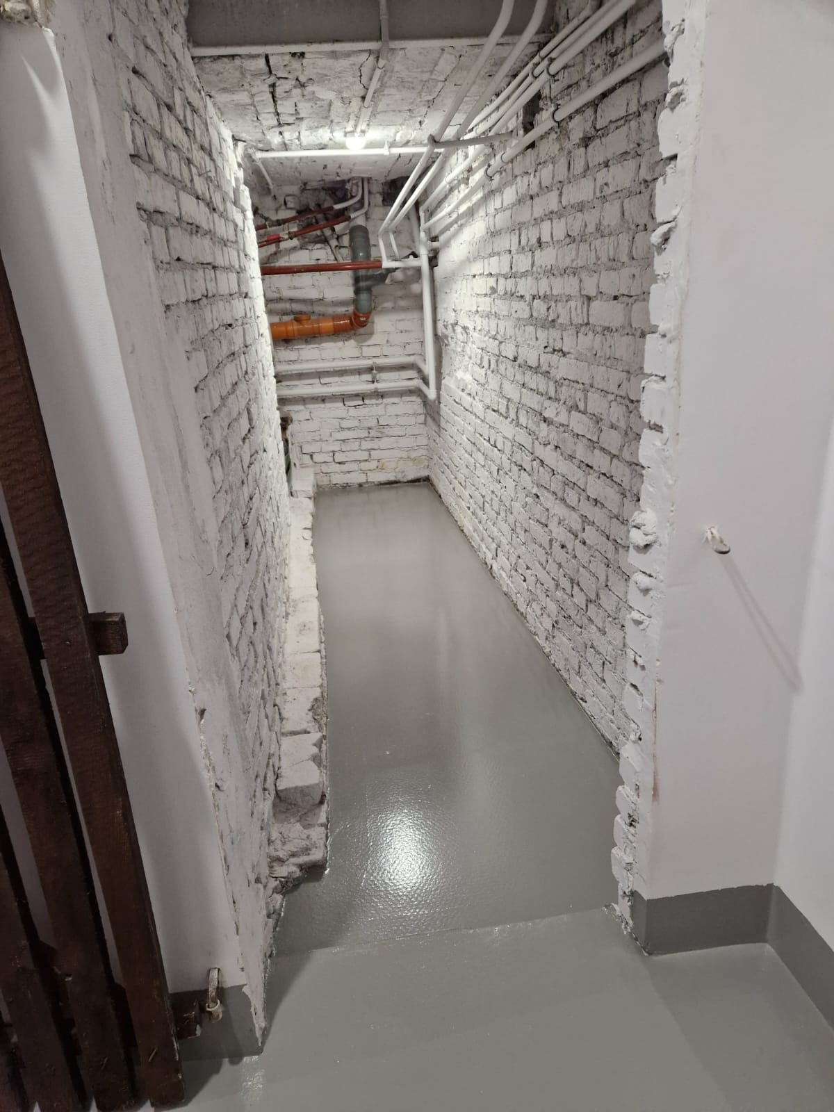 Pomieszczenie piwniczne z odnowioną, szarą posadzką, białe ceglane ściany z widoczną instalacją rur pod sufitem, widoczne fragmenty drewnianej konstrukcji z lewej strony.