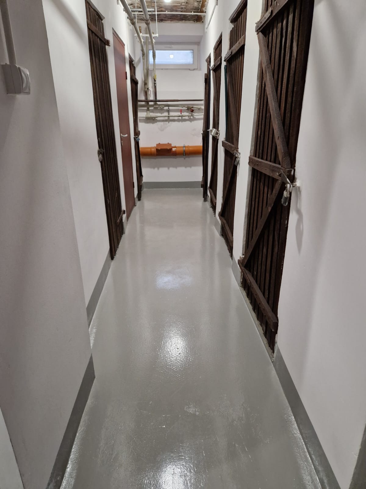 Korytarz z rzędem drewnianych boksów z kłódkami, szare ściany i podłoga z widoczną instalacją wodno-kanalizacyjną pod sufitem.