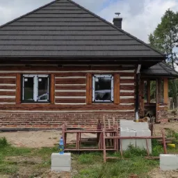 Dom z bali z nowymi oknami PCV w trakcie montażu, widoczne folie ochronne, rusztowanie i materiały budowlane na posesji.