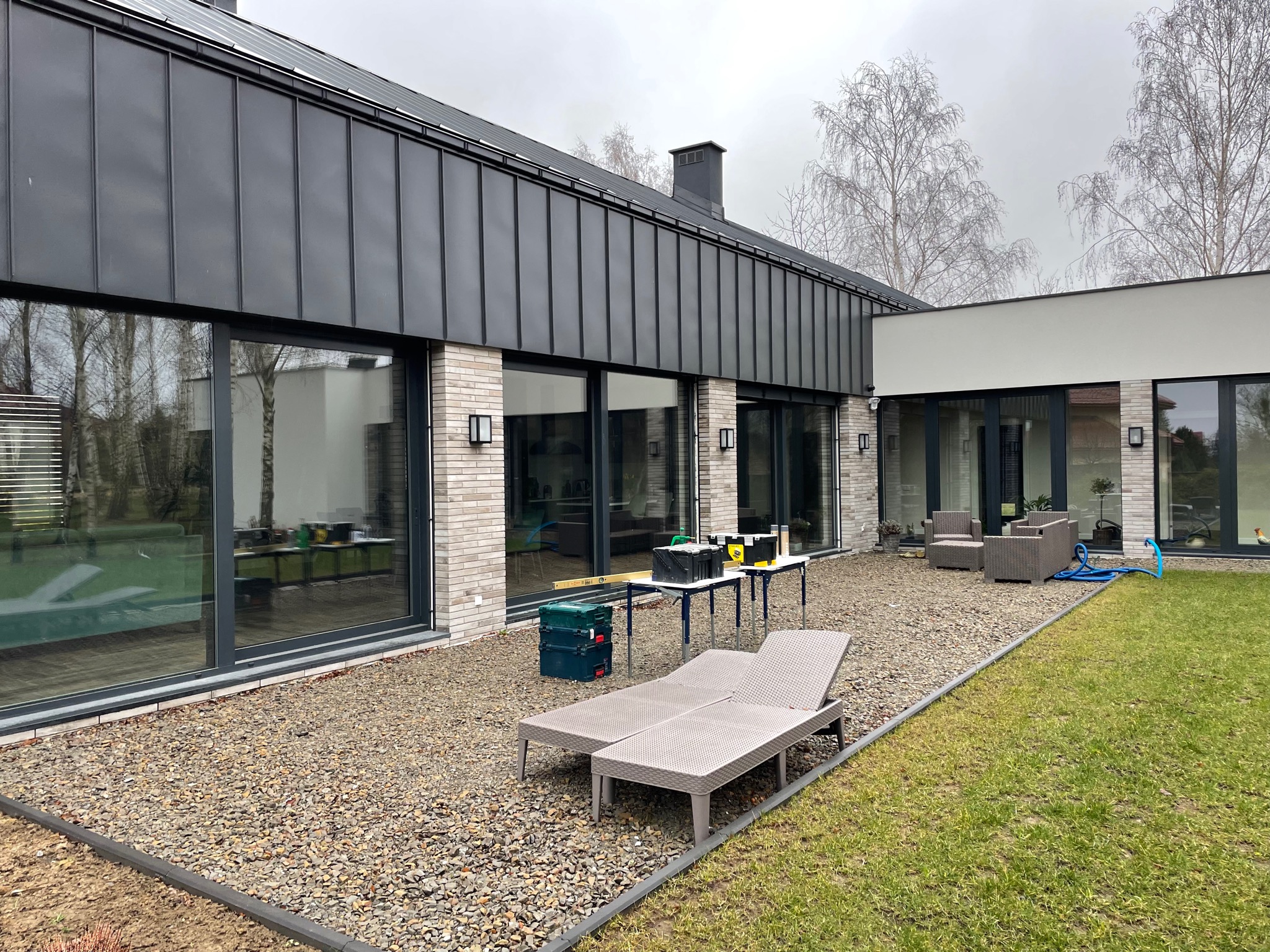 Nowoczesny dom z grafitową fasadą i dużymi oknami, przed którymi rozstawione są narzędzia i stolik warsztatowy, sugerujące prace montażowe lub serwisowe.