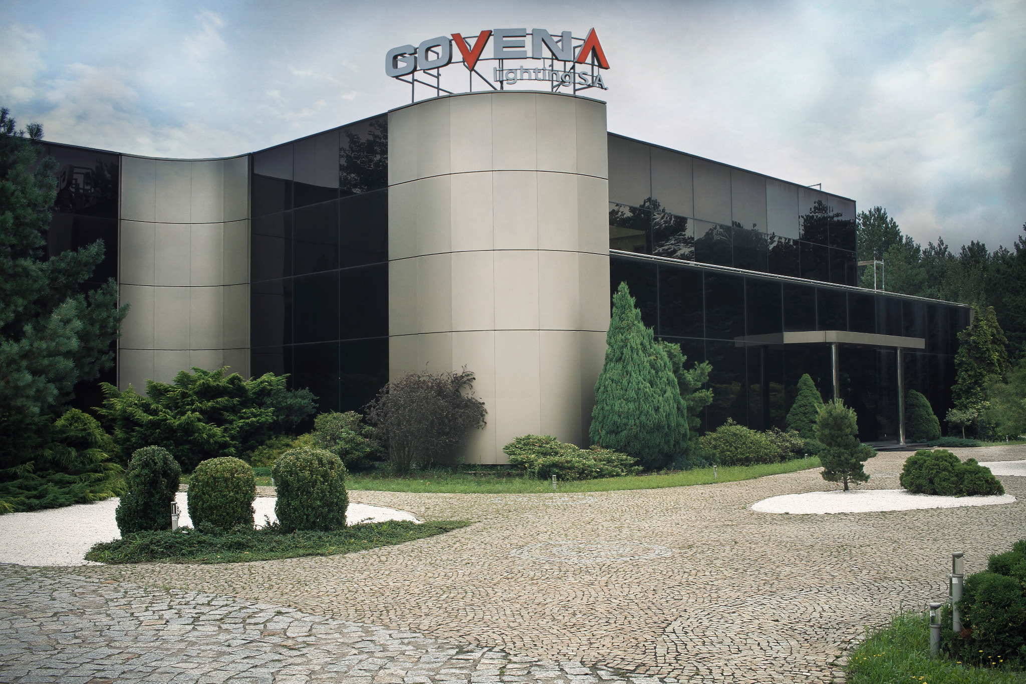 Nowoczesny budynek biurowy z ciemnymi oknami i zaokrągloną wieżą z logo firmy oświetleniowej na szczycie, otoczony zadbaną zielenią i brukowanym podjazdem.