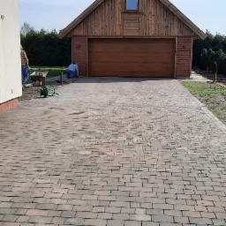 Ułożona kostka brukowa prowadząca do garażu z drewnianą bramą, widoczne dwie osoby pracujące, taczka z piaskiem i rząd tuj.