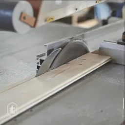 Cięcie deski z jasnego drewna za pomocą piły stołowej z aluminiową prowadnicą, widoczne drobne wióry.
