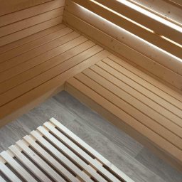 Tra.studio stolarstwo - Wnętrze sauny z drewnianymi ławkami, podestem i koszem wypełnionym kamieniami do nagrzewania.