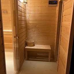 Tra.studio stolarstwo - Wnętrze sauny wykończone jasnym drewnem, z ławką, wiadrem i chochlą, oświetlone ciepłym światłem lampy ściennej.