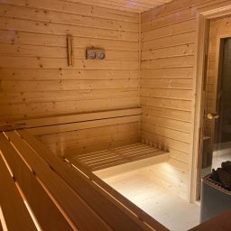 Tra.studio stolarstwo - Wnętrze drewnianej sauny z ławkami, piecem z kamieniami i szklanymi drzwiami, na ścianie termometr i klepsydra odmierzająca czas