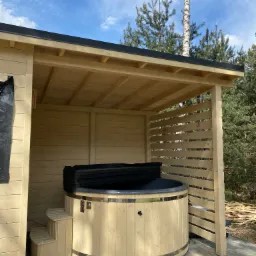 Drewniana balia z pokrywą pod zadaszeniem z ażurową ścianą, widoczne schodki wejściowe i fragmenty konstrukcji z desek w tle.