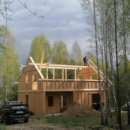Tra.studio stolarstwo - Konstrukcja dwukondygnacyjnego domu w stanie surowym otwartym, wykonana z drewnianych belek i płyt OSB, z widocznym pracownikiem na dachu, na tle brzozowego lasu i pochmurnego nieba. Na pierwszym...