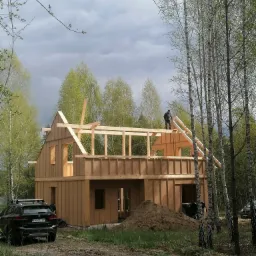 Konstrukcja dwukondygnacyjnego domu w stanie surowym otwartym, wykonana z drewnianych belek i płyt OSB, z widocznym pracownikiem na dachu, na tle brzozowego lasu i pochmurnego nieba. Na pierwszym...