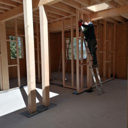 Tra.studio stolarstwo - Konstrukcja drewniana budynku w trakcie montażu, pracownik na drabinie mocuje elementy, widoczne okno i surowe podłoże.