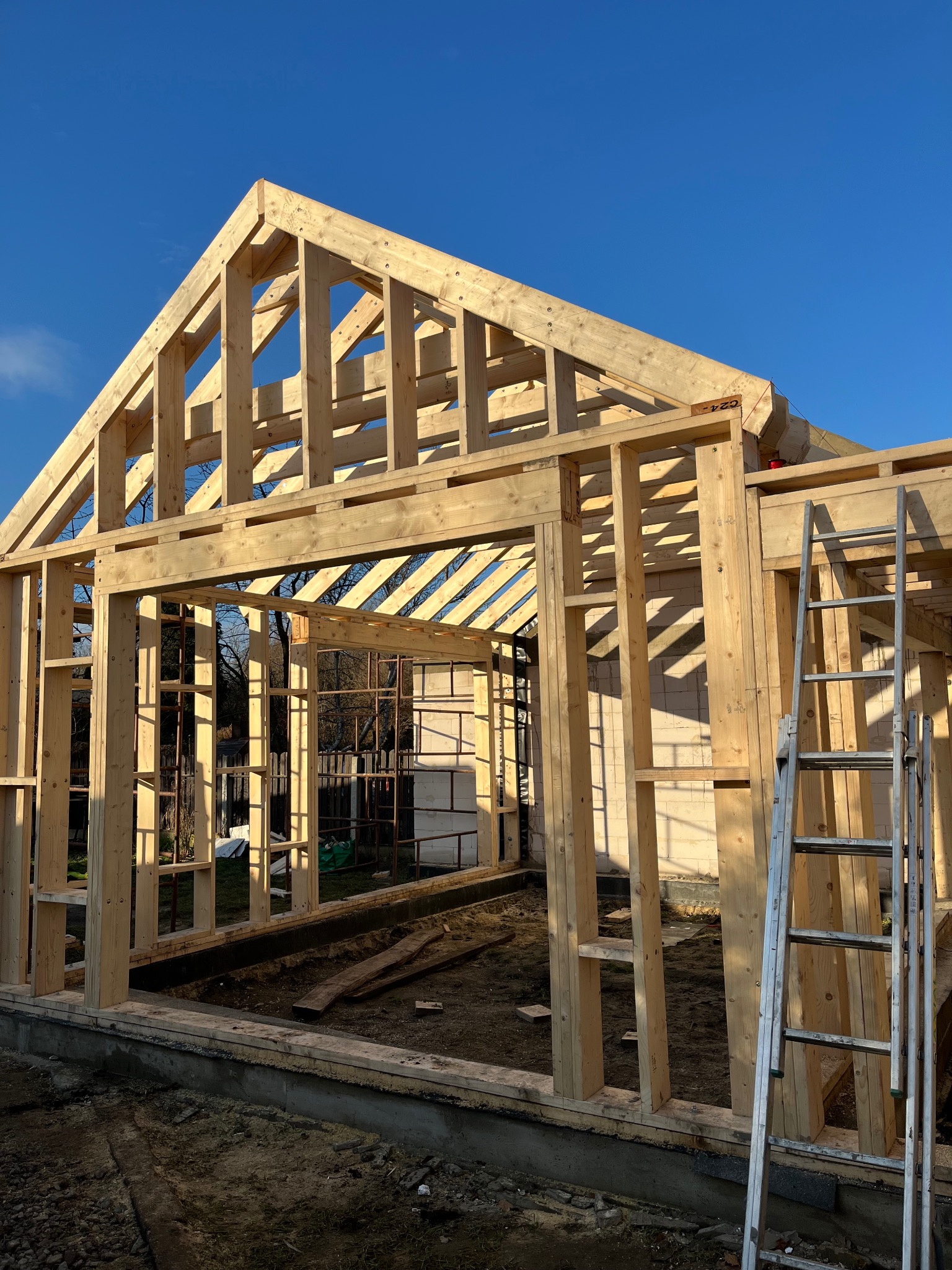Wznoszenie drewnianej konstrukcji budynku w słoneczny dzień, widoczna drabina oparta o ścianę, fundament oraz elementy konstrukcyjne z jasnego drewna na tle błękitnego nieba.
