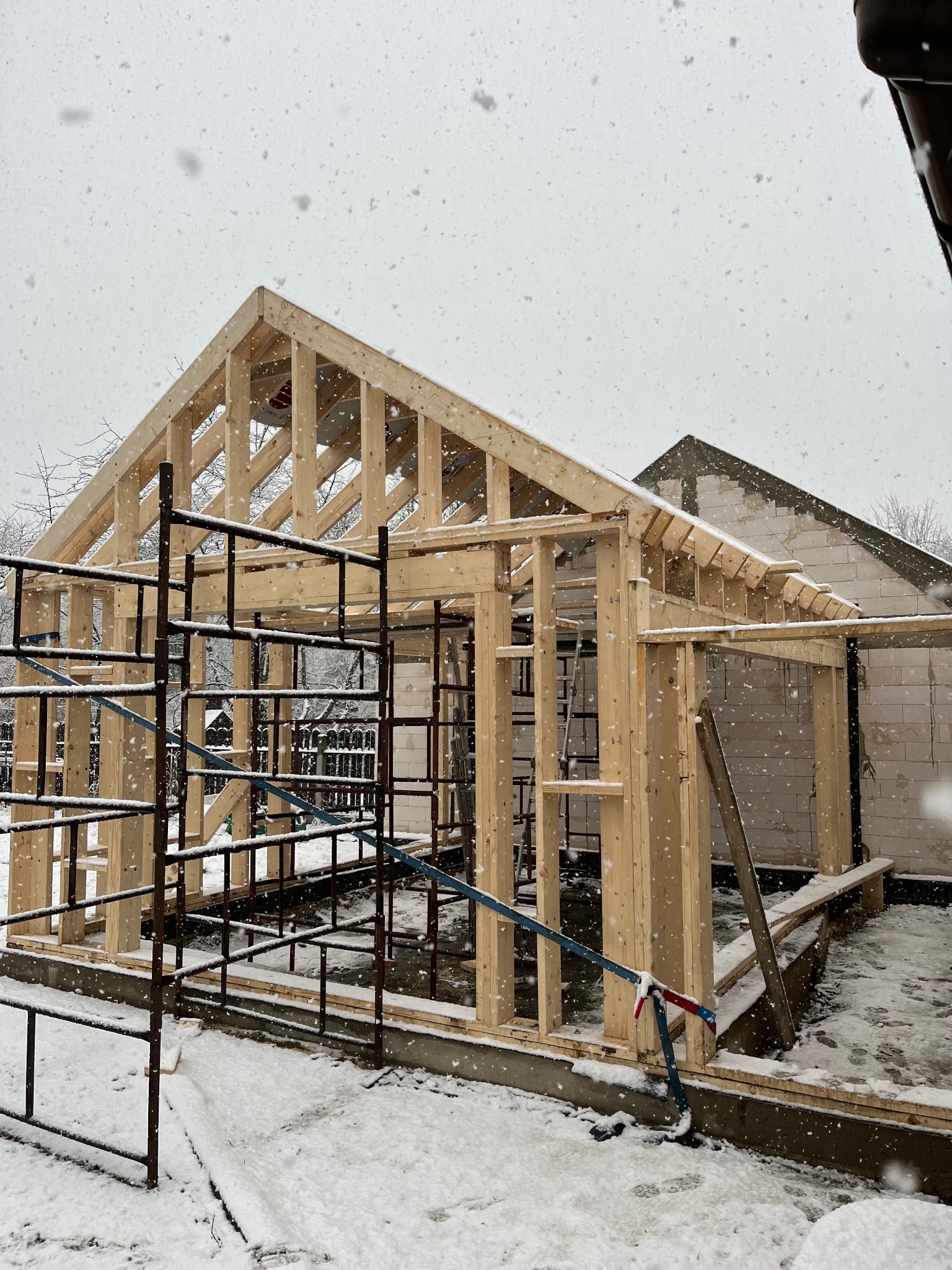 Konstrukcja domu w trakcie budowy, widoczny szkielet z drewnianych belek i metalowe rusztowanie, wszystko przyprószone śniegiem podczas zimowej pogody.