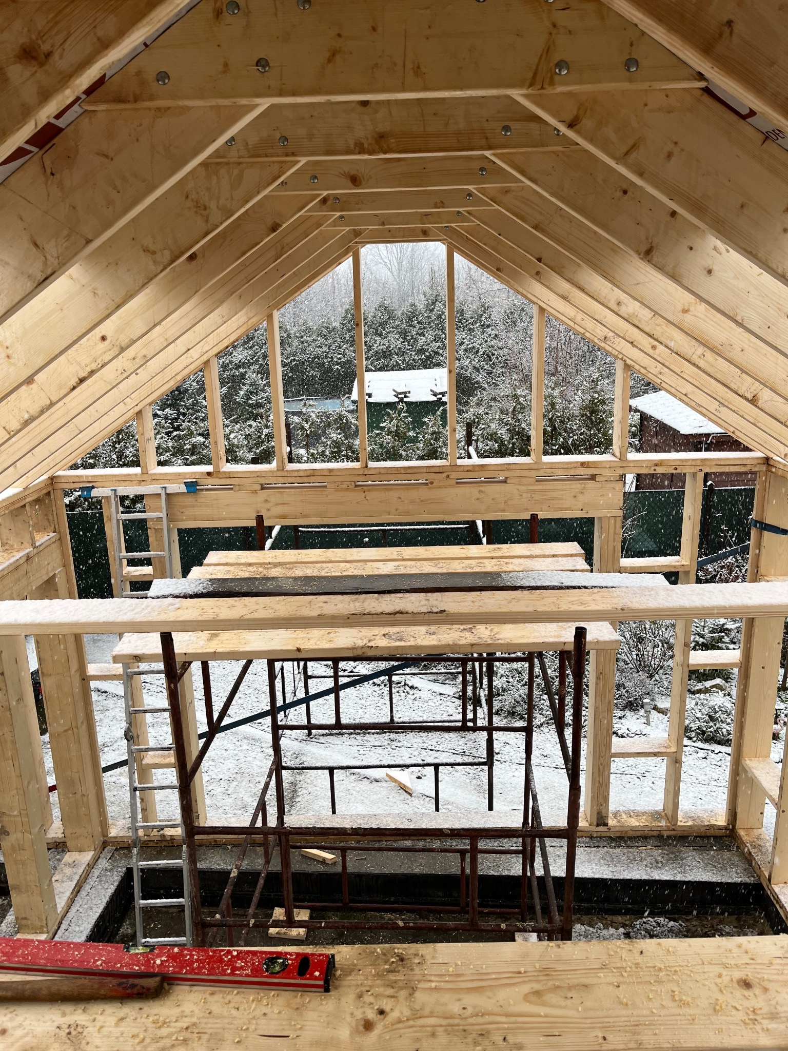 Wnętrze konstrukcji domu w trakcie budowy, widoczne drewniane elementy szkieletu, rusztowania, drabina i poziomica, na zewnątrz pada śnieg.