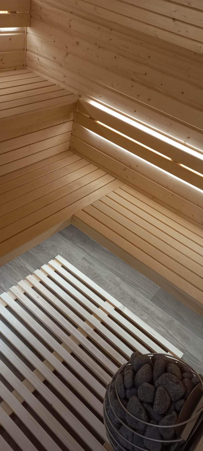 Wnętrze sauny z drewnianymi ławkami, podestem i koszem wypełnionym kamieniami do nagrzewania.