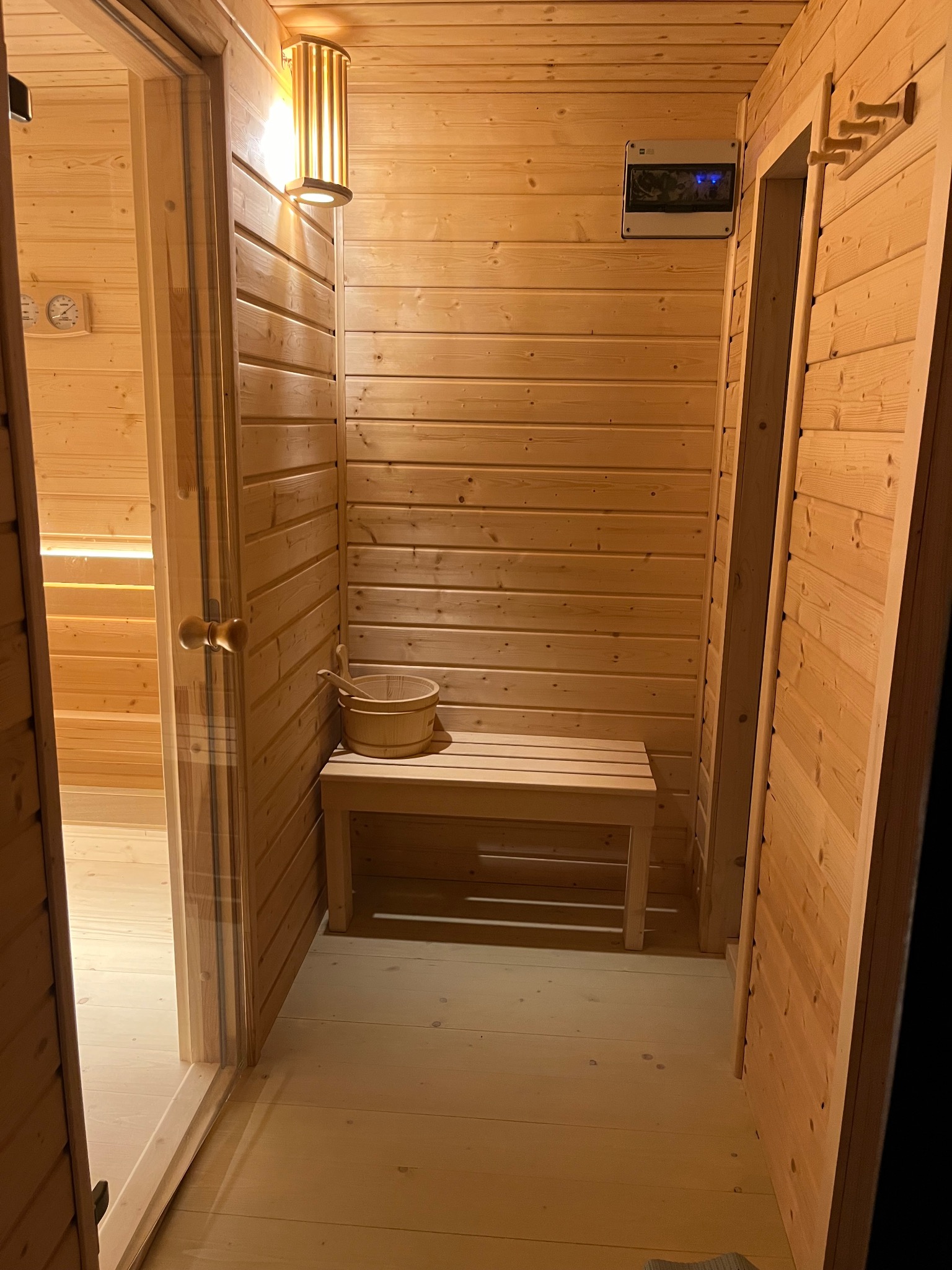 Wnętrze sauny wykończone jasnym drewnem, z ławką, wiadrem i chochlą, oświetlone ciepłym światłem lampy ściennej.