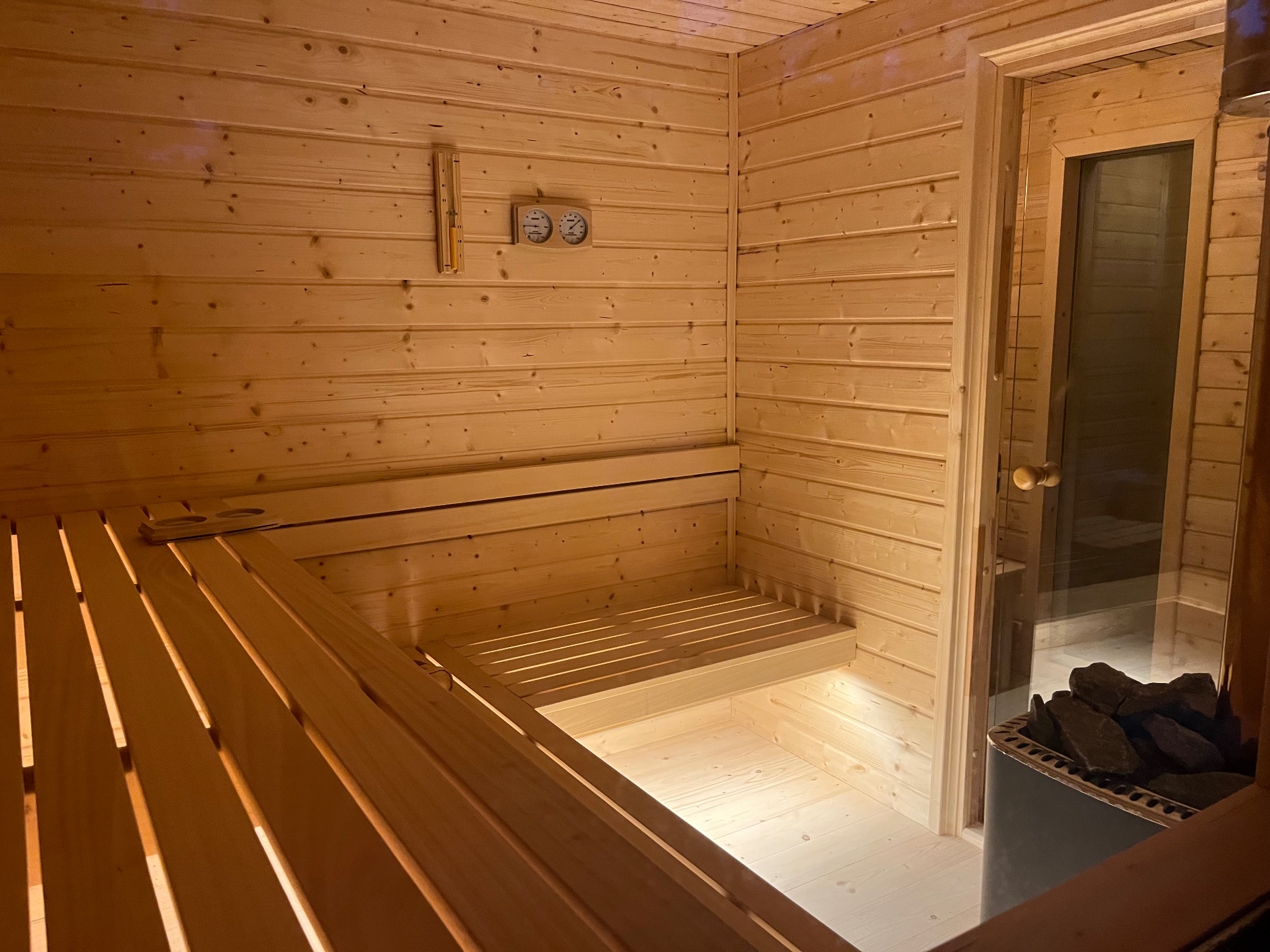 Wnętrze drewnianej sauny z ławkami, piecem z kamieniami i szklanymi drzwiami, na ścianie termometr i klepsydra odmierzająca czas