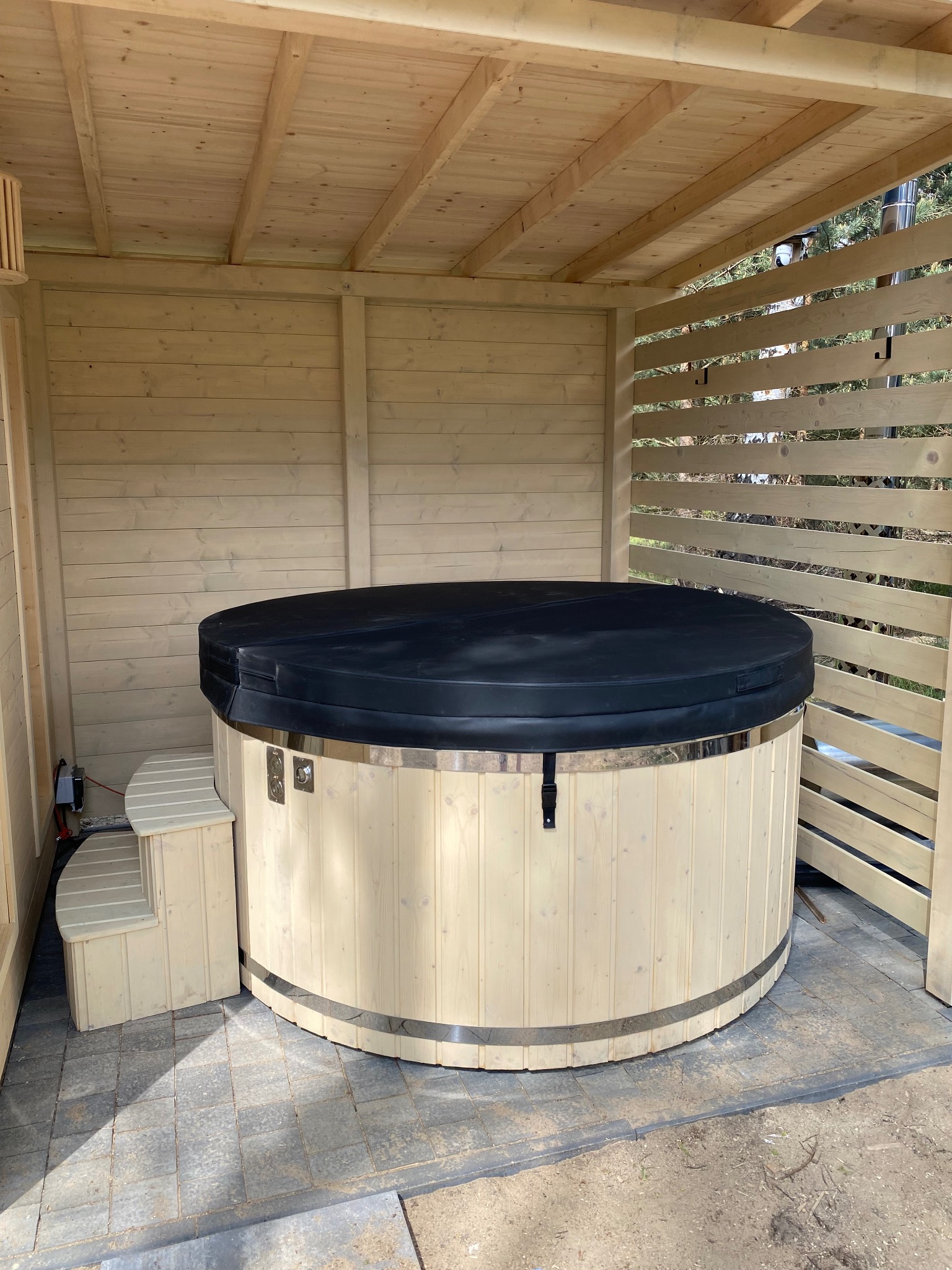 Okrągła, drewniana wanna z hydromasażem, przykryta czarną pokrywą, umieszczona w drewnianej konstrukcji z ażurowymi ścianami, z dołączonymi schodkami.