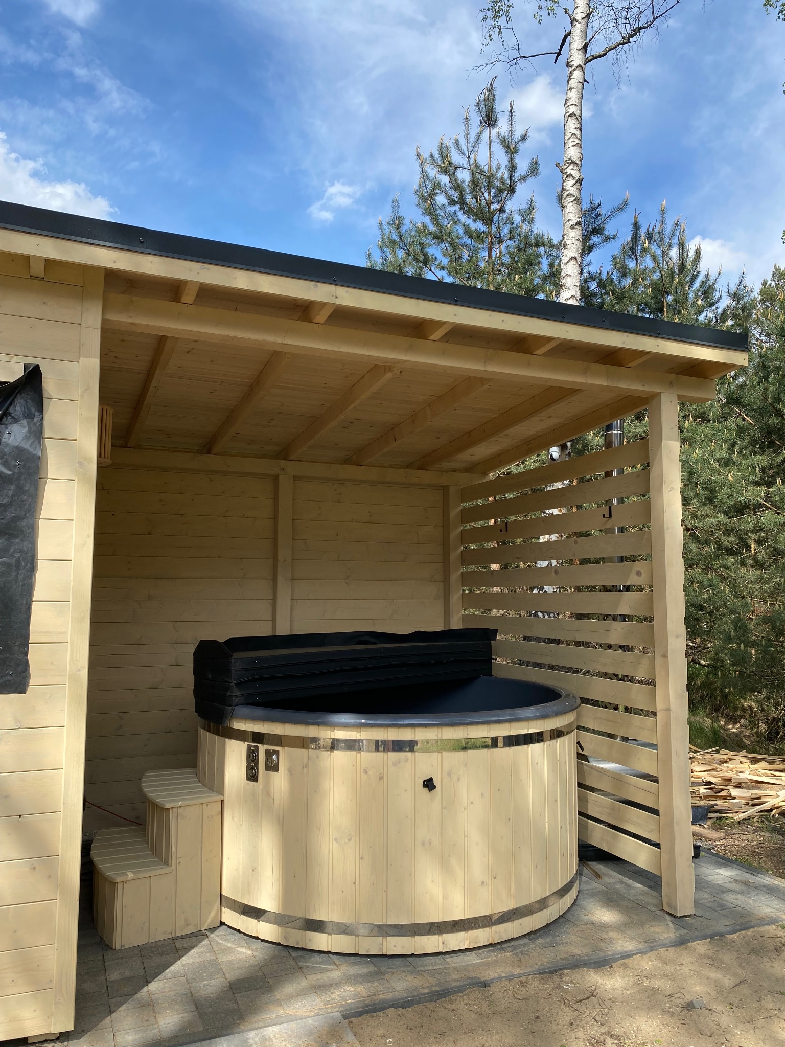 Drewniana balia z pokrywą pod zadaszeniem z ażurową ścianą, widoczne schodki wejściowe i fragmenty konstrukcji z desek w tle.