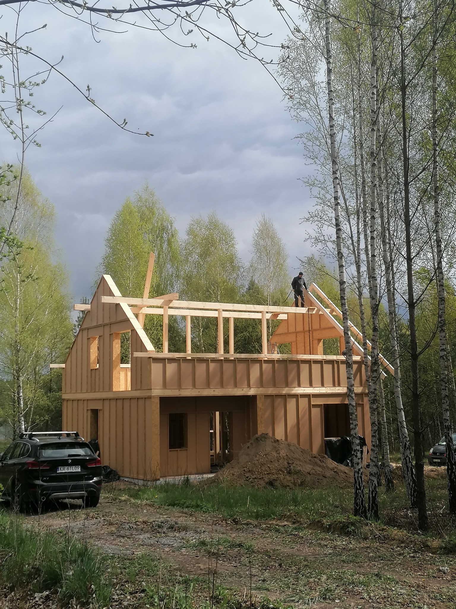 Konstrukcja dwukondygnacyjnego domu w stanie surowym otwartym, wykonana z drewnianych belek i płyt OSB, z widocznym pracownikiem na dachu, na tle brzozowego lasu i pochmurnego nieba. Na pierwszym...