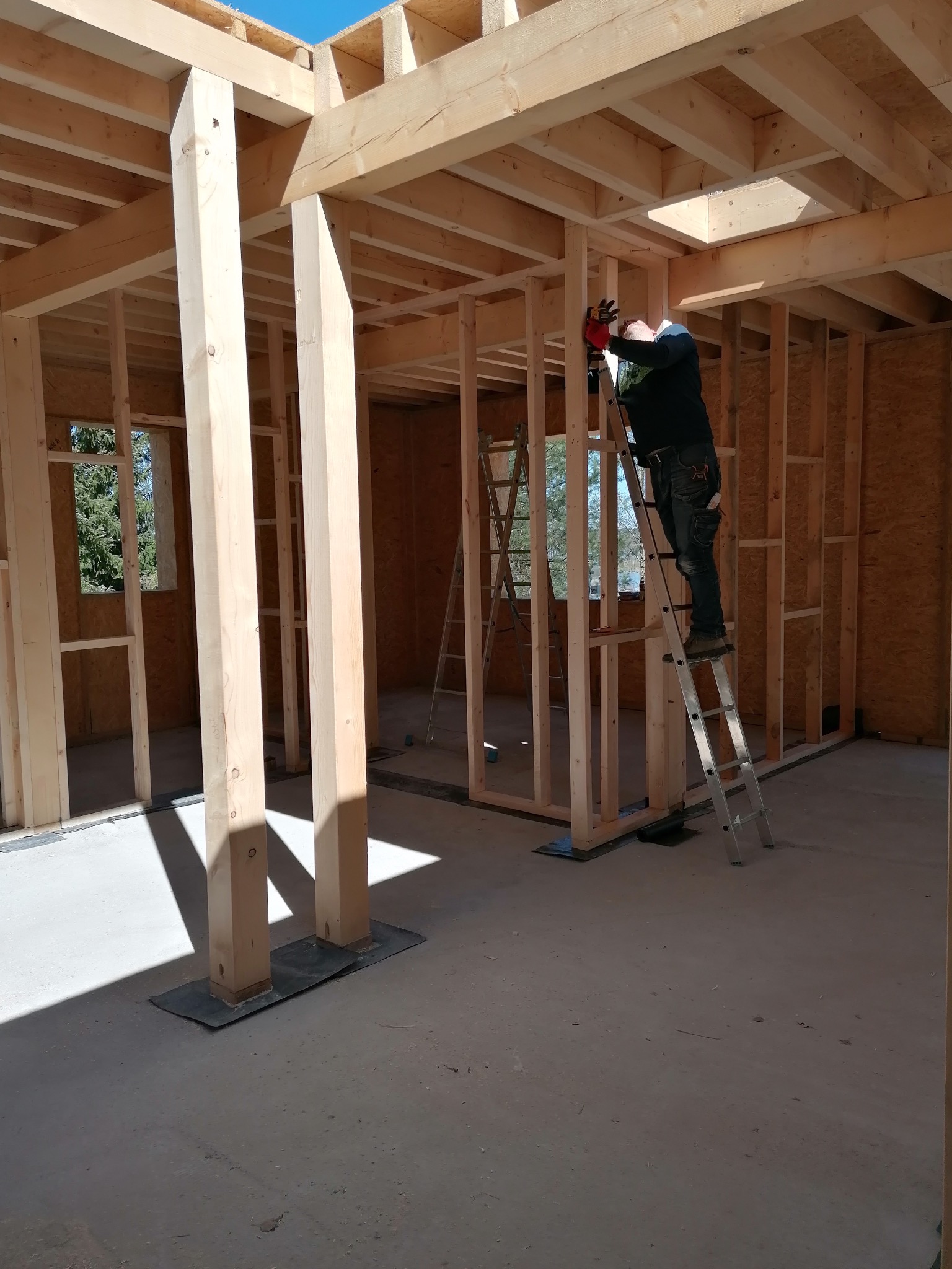 Konstrukcja drewniana budynku w trakcie montażu, pracownik na drabinie mocuje elementy, widoczne okno i surowe podłoże.