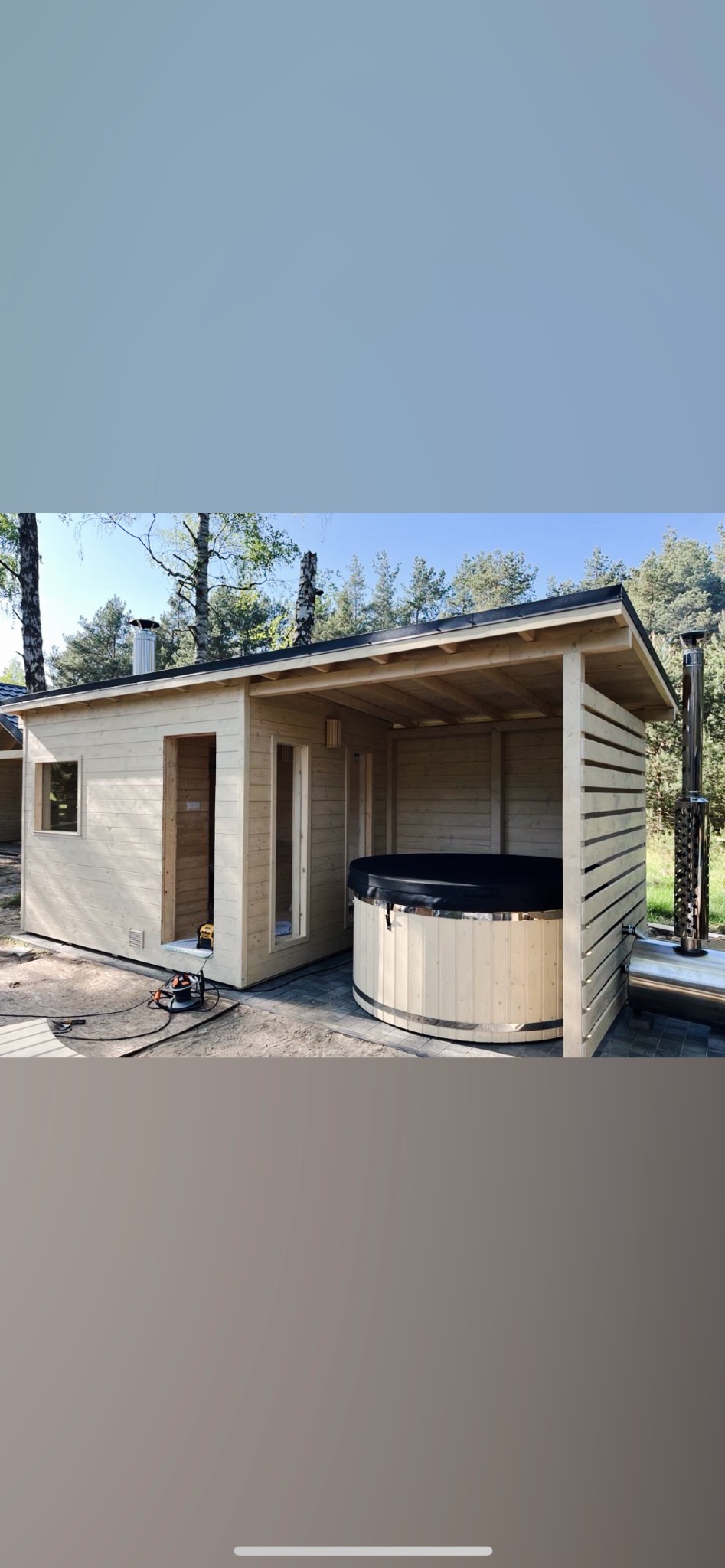 Drewniana konstrukcja z podcieniem kryjącym okrągłą balię z czarną pokrywą, obok widoczny komin, na pierwszym planie narzędzia sugerujące prace wykończeniowe.