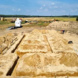 Wykonane wykopy pod fundamenty domu jednorodzinnego, widoczne drewniane szalunki, w tle biały bus firmy budowlanej i krajobraz polny w słoneczny dzień.