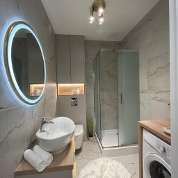 MA-KAL SP.Z.O.O. - Nowoczesna łazienka z marmurowymi płytkami, okrągłym lustrem z podświetleniem LED, kabiną prysznicową i pralką. Umywalka nablatowa z ręcznikami i dekoracyjne oświetlenie.