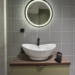 MA-KAL SP.Z.O.O. - Biała umywalka nablatowa z czarną armaturą na tle ściany z wzorzystych, czarno-białych płytek i okrągłego lustra z podświetleniem LED w nowej łazience.