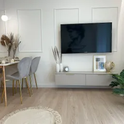 Jasne wnętrze salonu z drewnianą podłogą, szarymi krzesłami z złotymi nogami i telewizorem z wizerunkiem rzeźby Dawida na ścianie. Minimalistyczny design.