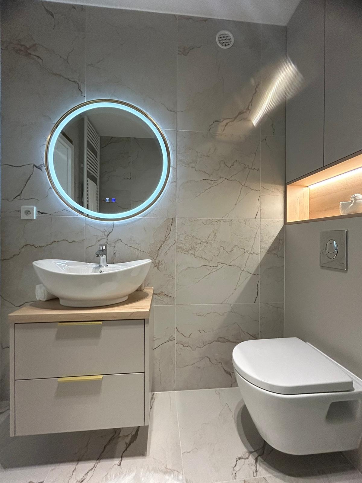 Jasna łazienka z marmurowymi płytkami, okrągłym lustrem LED, wiszącą toaletą i szafką pod umywalkę z drewnianym blatem i złotymi uchwytami.