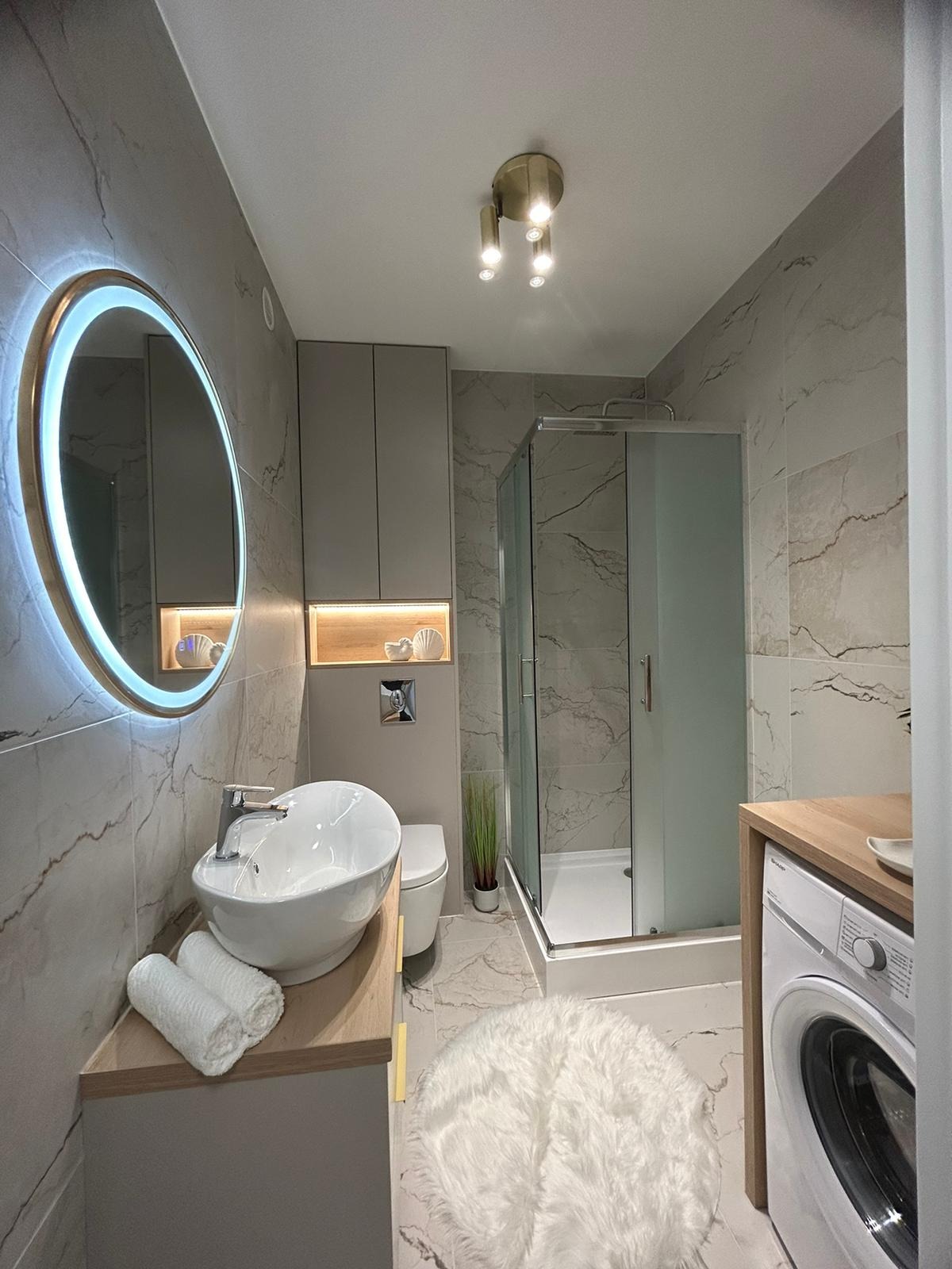 Nowoczesna łazienka z marmurowymi płytkami, okrągłym lustrem z podświetleniem LED, kabiną prysznicową i pralką. Umywalka nablatowa z ręcznikami i dekoracyjne oświetlenie.