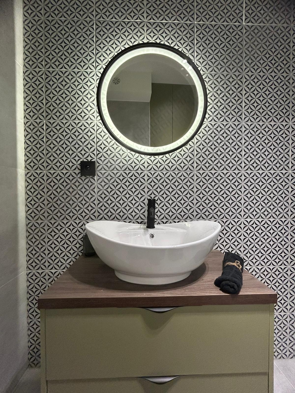 Biała umywalka nablatowa z czarną armaturą na tle ściany z wzorzystych, czarno-białych płytek i okrągłego lustra z podświetleniem LED w nowej łazience.