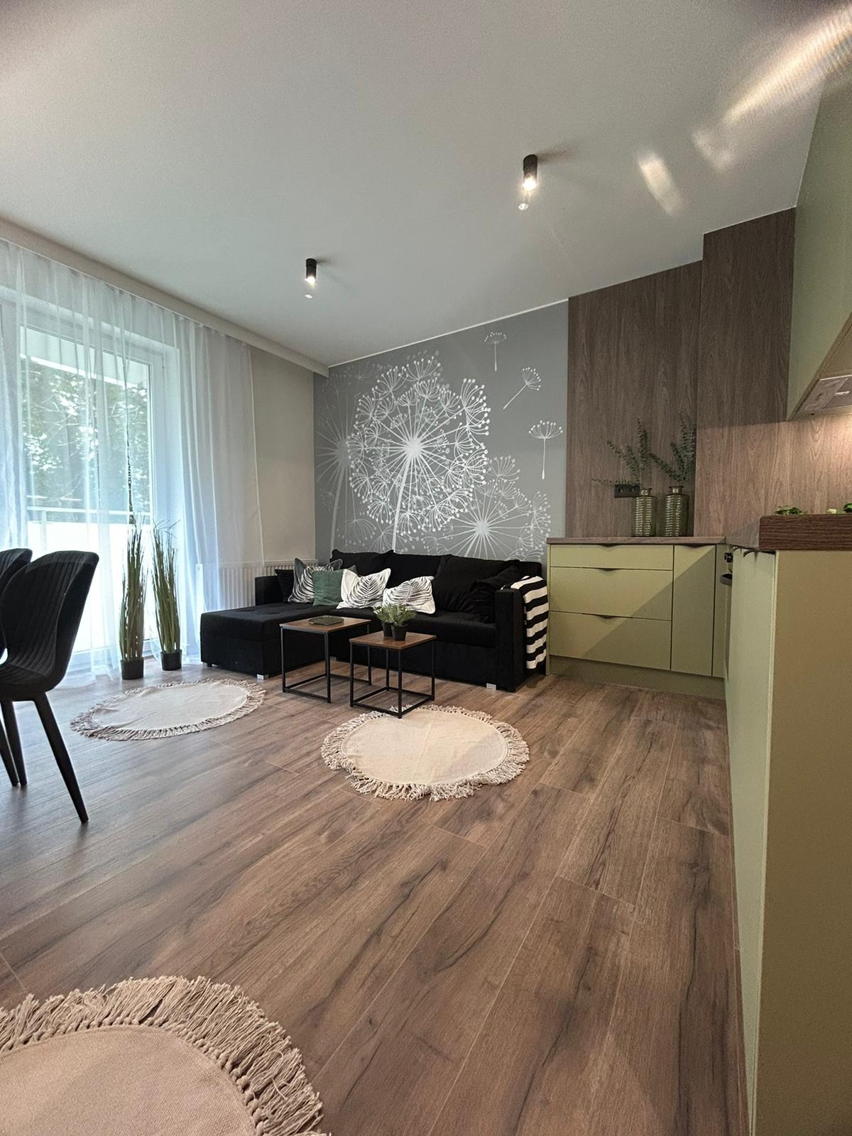 Salon z drewnopodobną podłogą, czarną sofą i ścianą z motywem dmuchawca. Widoczne okno z białymi zasłonami i fragment kuchni w odcieniach zieleni.
